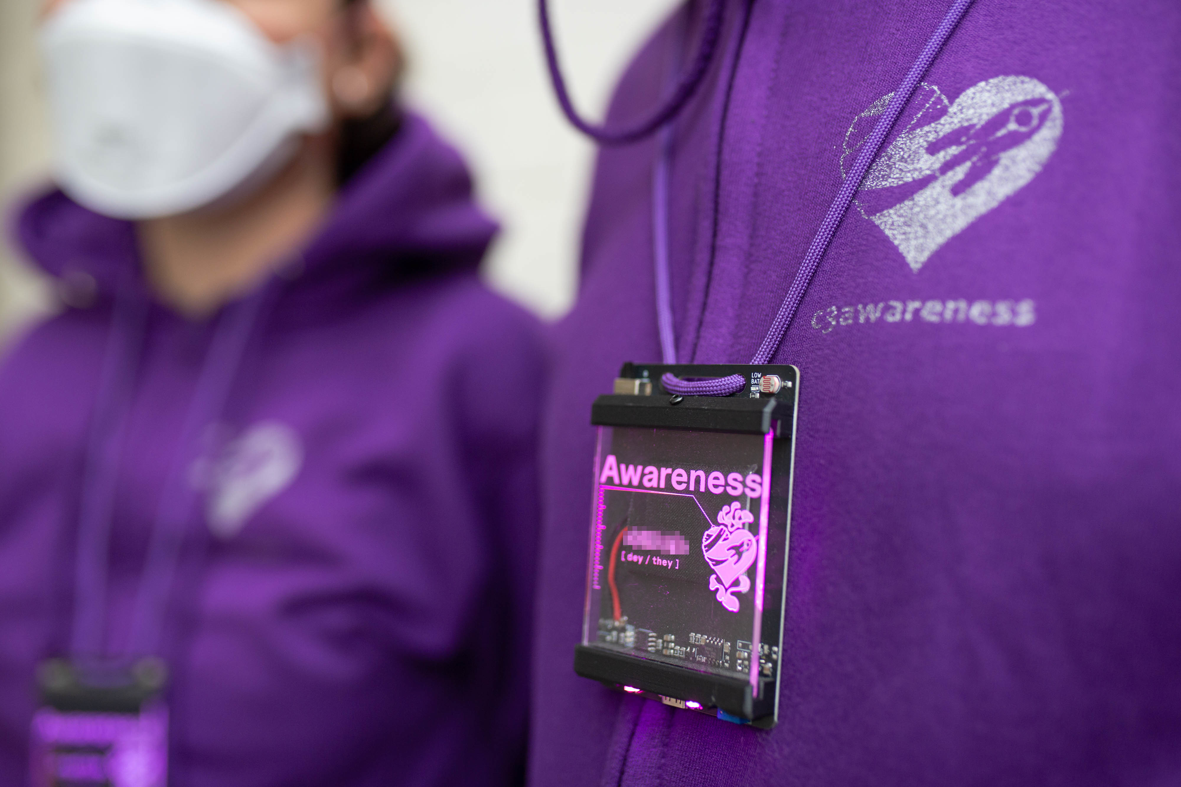 Auf dem Foto sind die Oberkörper zweier Personen zu erkennen. Sie tragen lila Hoodies und lila leuchten Elektronik-Badges. Auf Hoodie und Badge ist das c3awareness-Logo zu erkennen: Ein Herz um das eine Rakete eine Bandage spannt. Auf der Badge leuchtet die Überschrift 'Awareness', darunter steht ein verpixelter Name sowie Pronomen.  Photo (c) c3awareness | TRim